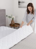 Kobieta układa biały ochraniacz na łóżku w jasnym wnętrzu z kwiatami i stolikiem obok