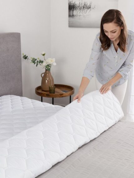 Kobieta układa biały ochraniacz na łóżku w jasnym wnętrzu z kwiatami i stolikiem obok