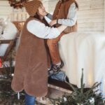 Mama w brązowej kamizelce i dziecko w stajni z choinkami i kocem