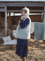 Kobieta w szarej kamizelce z kapturem stoi na tle stodoły, w otoczeniu siana i koców