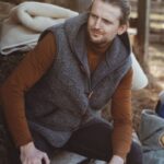 Mężczyzna w szarym kamizelce wełnianej siedzi na drewnianym podeście obok koców