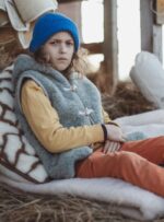 Dziecko w szarej kamizelce siedzi na kocu z wełny, oparty na poduszce w otoczeniu siana