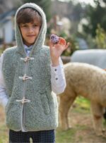 Chłopiec w szałwiowej kamizelce z kapturem trzyma ozdobione jajko, w tle alpaki na łące
