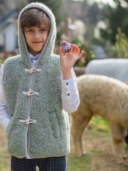 Chłopiec w szałwiowej kamizelce z kapturem trzyma ozdobione jajko, w tle alpaki na łące