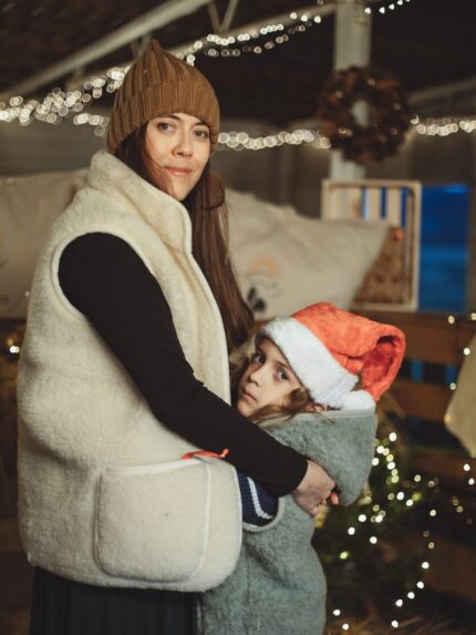 Kobieta w beżowej kamizelce trzyma dziecko w szałwiowej kamizelce z czerwoną czapką na tle świątecznych dekoracji