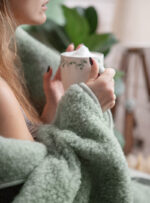 Kobieta owinięta zielonym kocem trzyma filiżankę w dłoniach w przytulnym wnętrzu