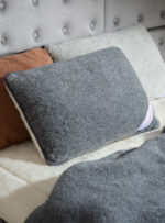 Grafitowa poduszka na tle jasnego łóżka z innymi poduszkami w odcieniach brązu i grafitu