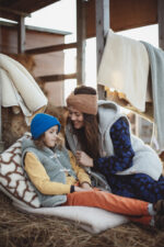 Kobieta z długimi włosami i dziecko w czapce siedzą na poduszce w stajni, wokół koce i siano. Oboje w wełnianych kamizelkach.