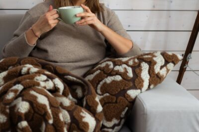 Kobieta w beżowym swetrze trzyma filiżankę na kanapie owinięta brązowym kocem, obok poduszka