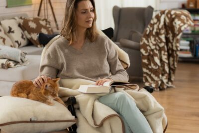Kobieta siedząca w fotelu z książką, obok niej leży kot, w tle przytulne wnętrze