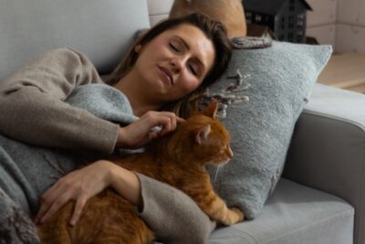 Kobieta na kanapie przytula kota, otulona szarym kocem, pod głową szara poduszka wełniana