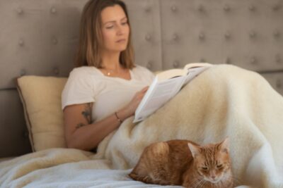 Kobieta czyta książkę na łóżku przykryta jasną kołdrą z wełny, obok leży rudy kot