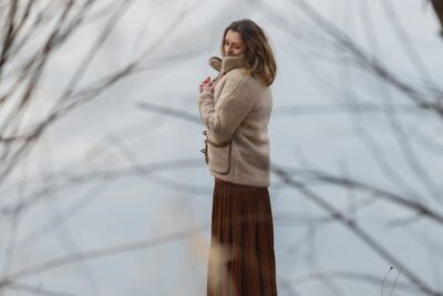 Kobieta w beżowej kurtce wełnianej i brązowej spódnicy stoi nad wodą, otoczona gałęziami