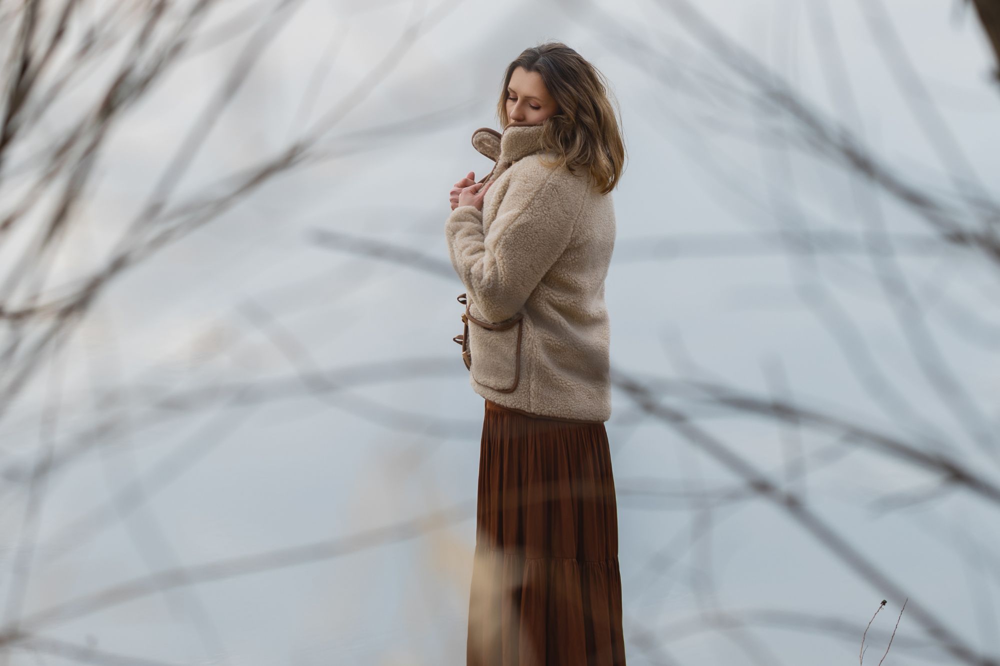 Kobieta w beżowej kurtce wełnianej i brązowej spódnicy stoi nad wodą, otoczona gałęziami