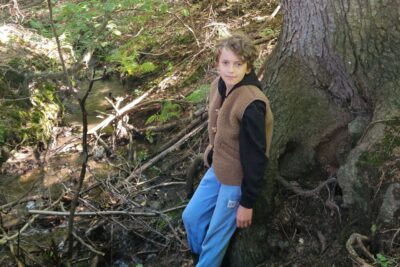 Chłopiec w brązowej kamizelce stoi przy strumieniu w lesie, w tle drzewa i gałęzie