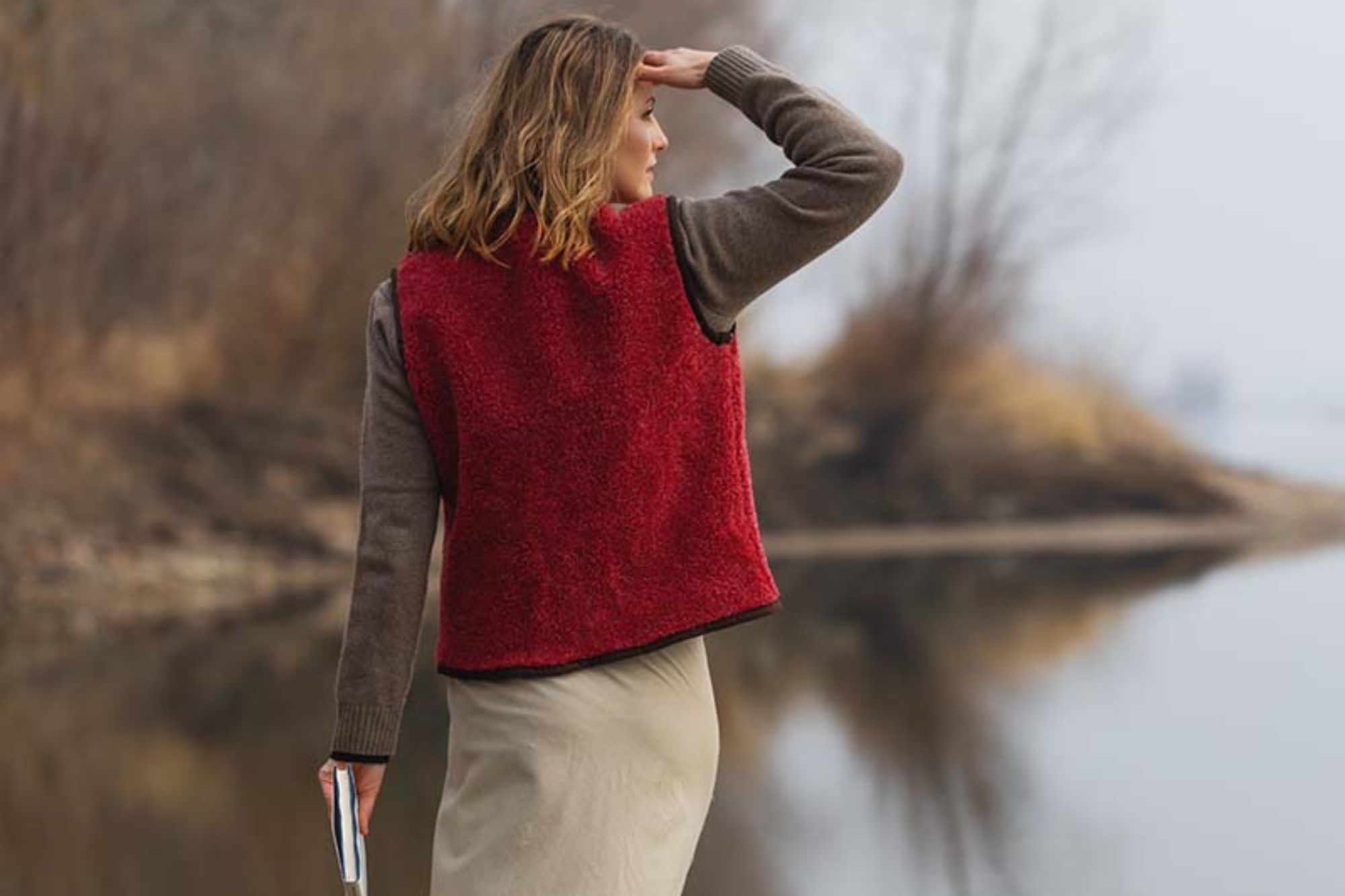 Kobieta w czerwonej kamizelce z wełny i brązowej bluzie stoi nad wodą, trzymając książkę