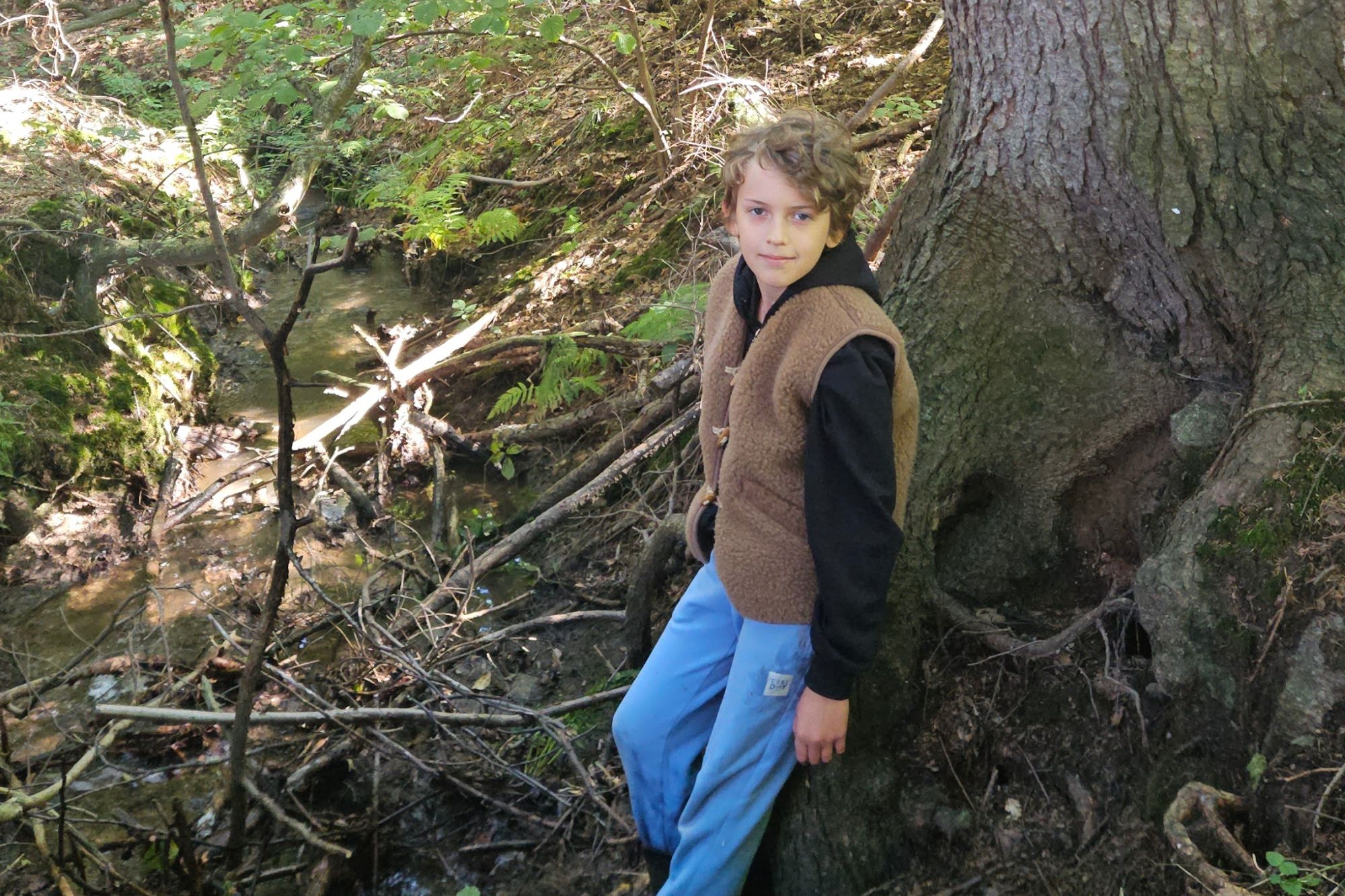 Chłopiec w brązowej kamizelce stoi przy strumieniu w lesie, w tle drzewa i gałęzie