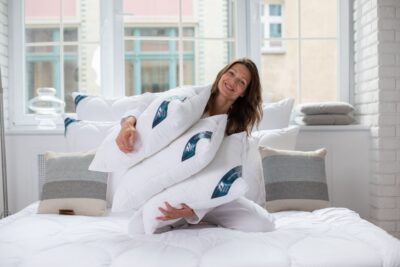 Kobieta siedząca na łóżku z białymi poduszkami, w tle jasne wnętrze z oknami