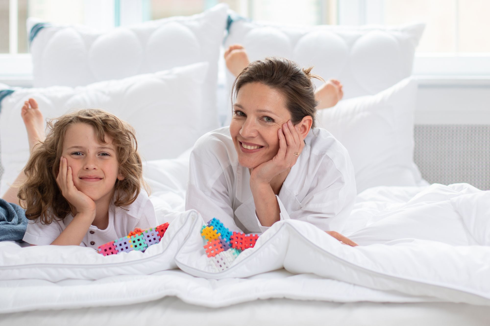 Kobieta i dziecko leżą na białej pościeli w jasnym pomieszczeniu, otoczeni poduszkami. Bawią się klockami