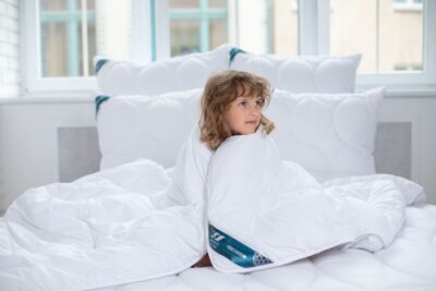 Chłopiec owinięty w białą kołdrę na łóżku w jasnym wnętrzu z poduszkami w tle