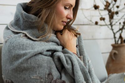 Kobieta owinięta szarym kocem, siedząca w przytulnym wnętrzu z roślinami w tle