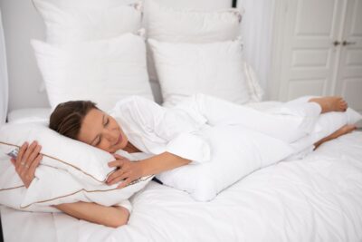 Kobieta leży na łóżku, przytulając poduszki, w otoczeniu białych pościeli