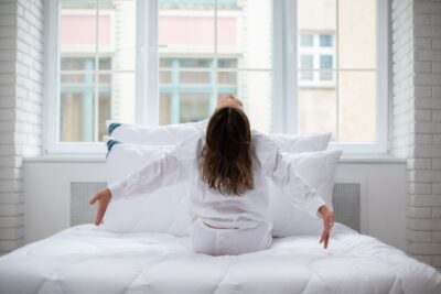 Kobieta w białej piżamie siedzi na białym łóżku z poduszkami w jasnym wnętrzu przy oknie