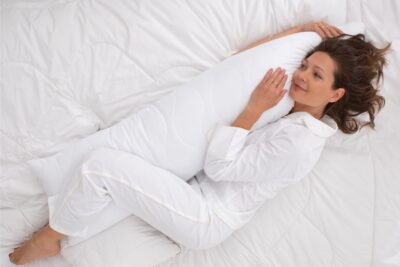 Kobieta w białej piżamie leży na łóżku i przytula poduszkę podłużną