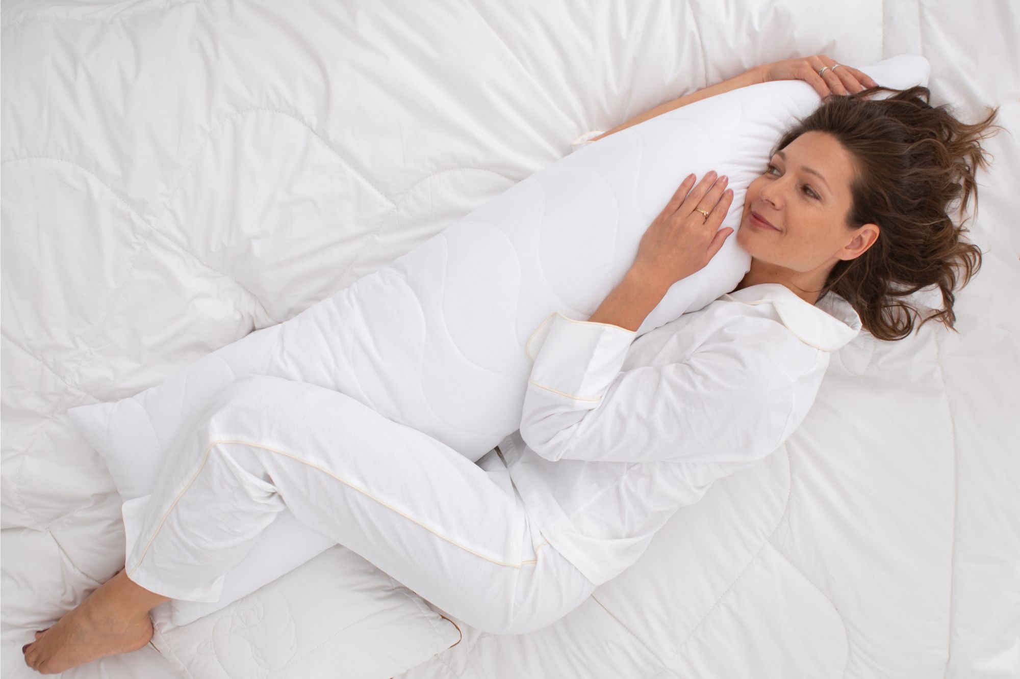 Kobieta w białej piżamie leży na łóżku i przytula poduszkę podłużną