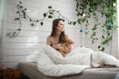 Kobieta siedząca na łóżku w białek kołdrze. Trzyma rudego kota. Rośliny na ścianie w tle