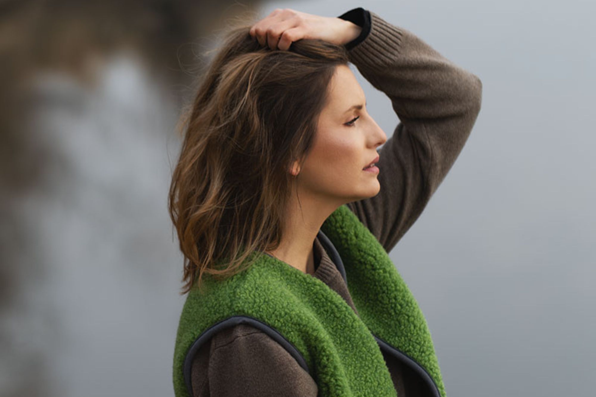 Kobieta w zielonej kamizelce z wełny, stojąca nad wodą, dotykająca włosów