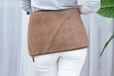 brązowy pas lędźwiowy na plecach kobiety w białych spodniach i szarym swetrze, w tle zasłony