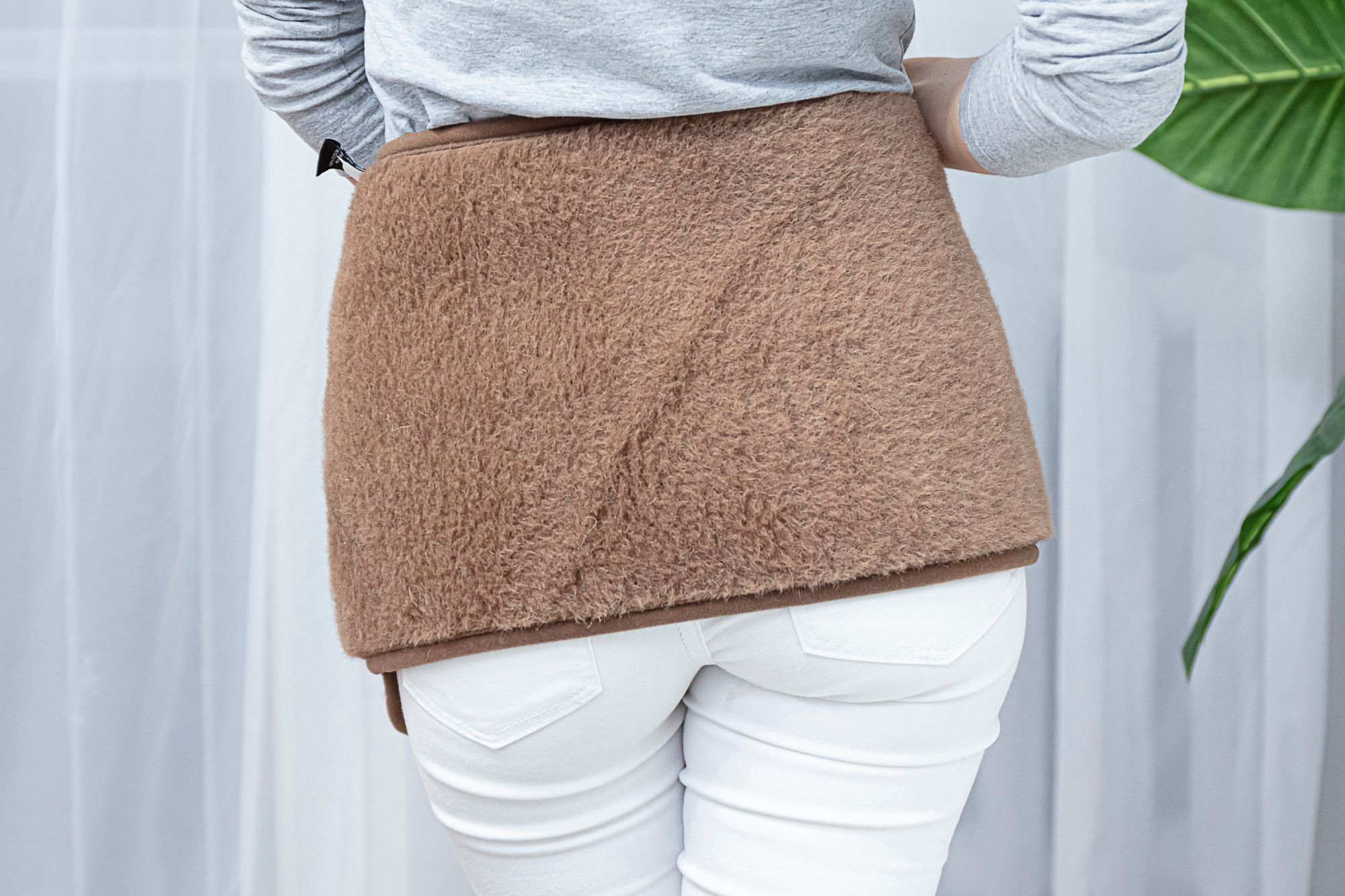 brązowy pas lędźwiowy na plecach kobiety w białych spodniach i szarym swetrze, w tle zasłony