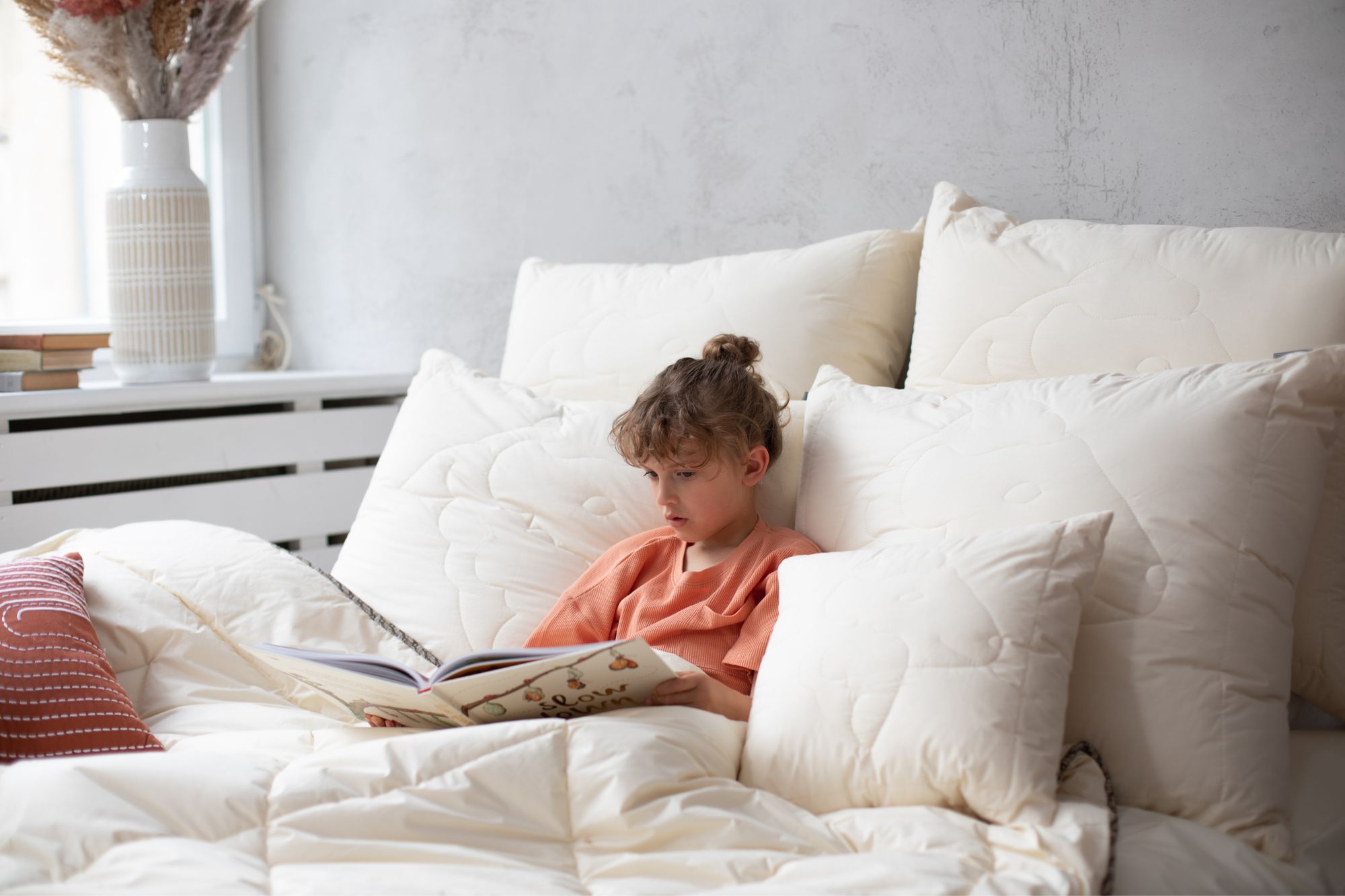 Chłopiec w pomarańczowej bluzce siedzi na łóżku otoczony poduszkami i czyta książkę
