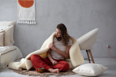 Kobieta owinięta jasnym kocem siedzi na podłodze w pokoju z poduszką i kubkiem