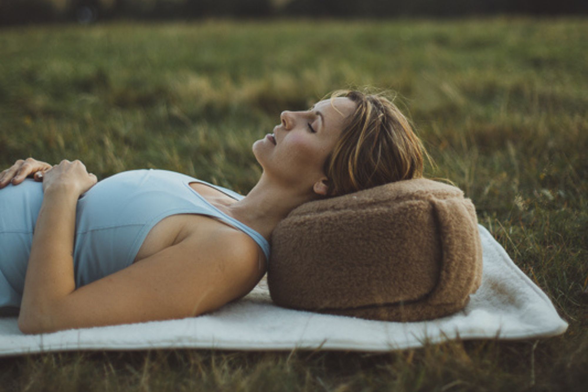 Kobieta w niebieskiej bluzce leży trawie na na macie do jogi z brązową poduszką do jogi pod głową