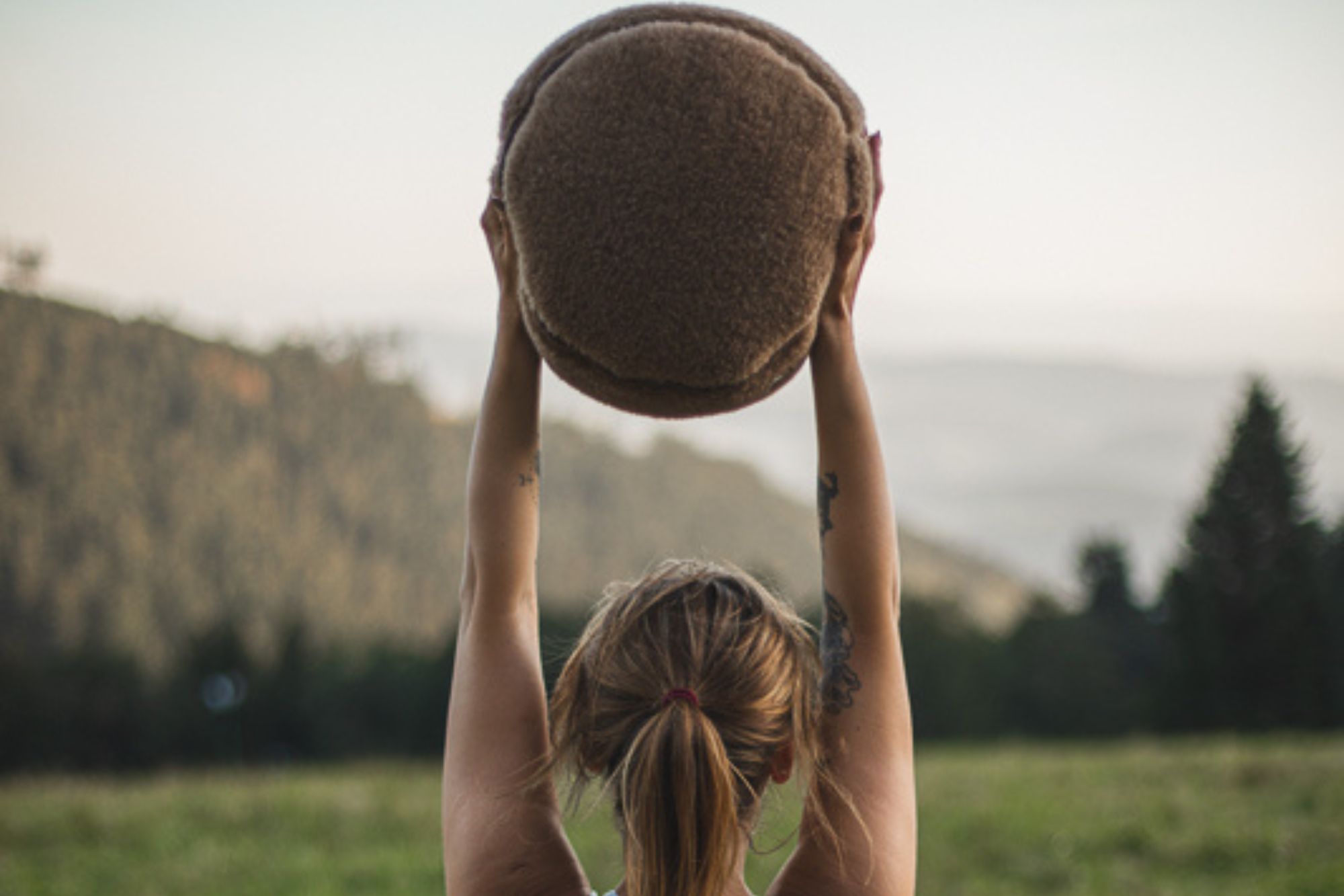 Kobieta trzyma w górze okrągłą poduszkę do medytacji w kolorze brązowym na tle górskiego krajobrazu