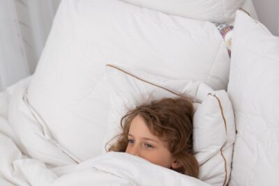 Chłopiec z brązowymi włosami owinięty w białą kołdrę leży na łóżku w sypialni