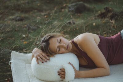 Kobieta w fioletowej bluzce leży na trawie, przytulając białą poduszkę do medytacji