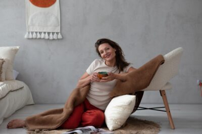 Kobieta siedząca na podłodze, owinięta w brązowy koc, trzyma kubek, w tle łóżko i krzesło