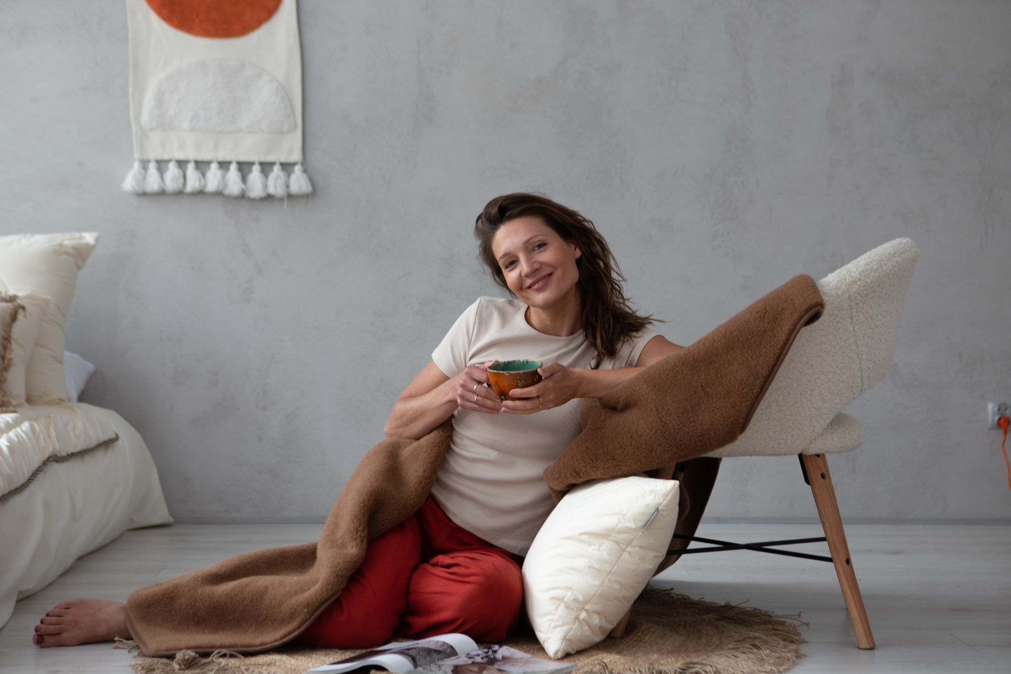 Kobieta siedząca na podłodze, owinięta w brązowy koc, trzyma kubek, w tle łóżko i krzesło