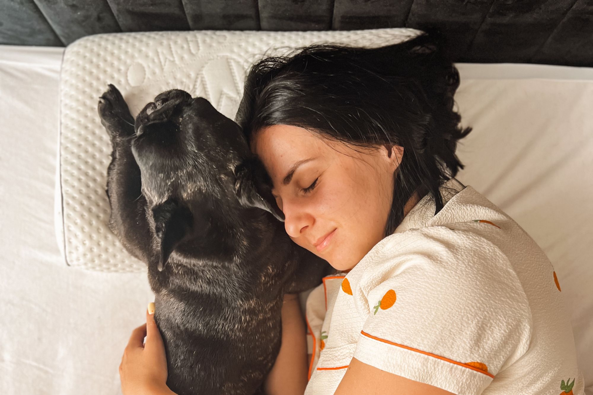 Kobieta leży na łóżku z zamkniętymi oczami, przytulając psa obok siebie. Leżą na poduszce ortopedycznej