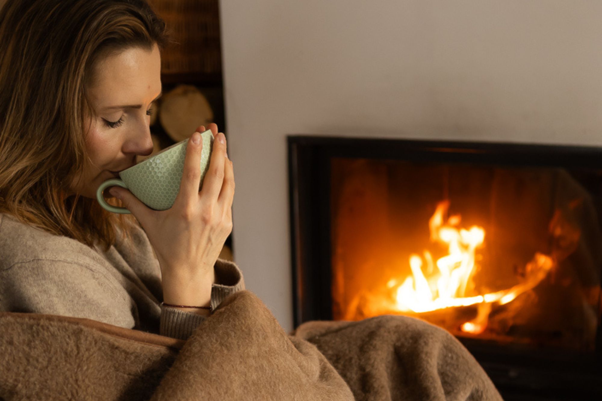 Kobieta w brązowym kocu trzyma zielony kubek, siedząc przy rozpalonym kominku