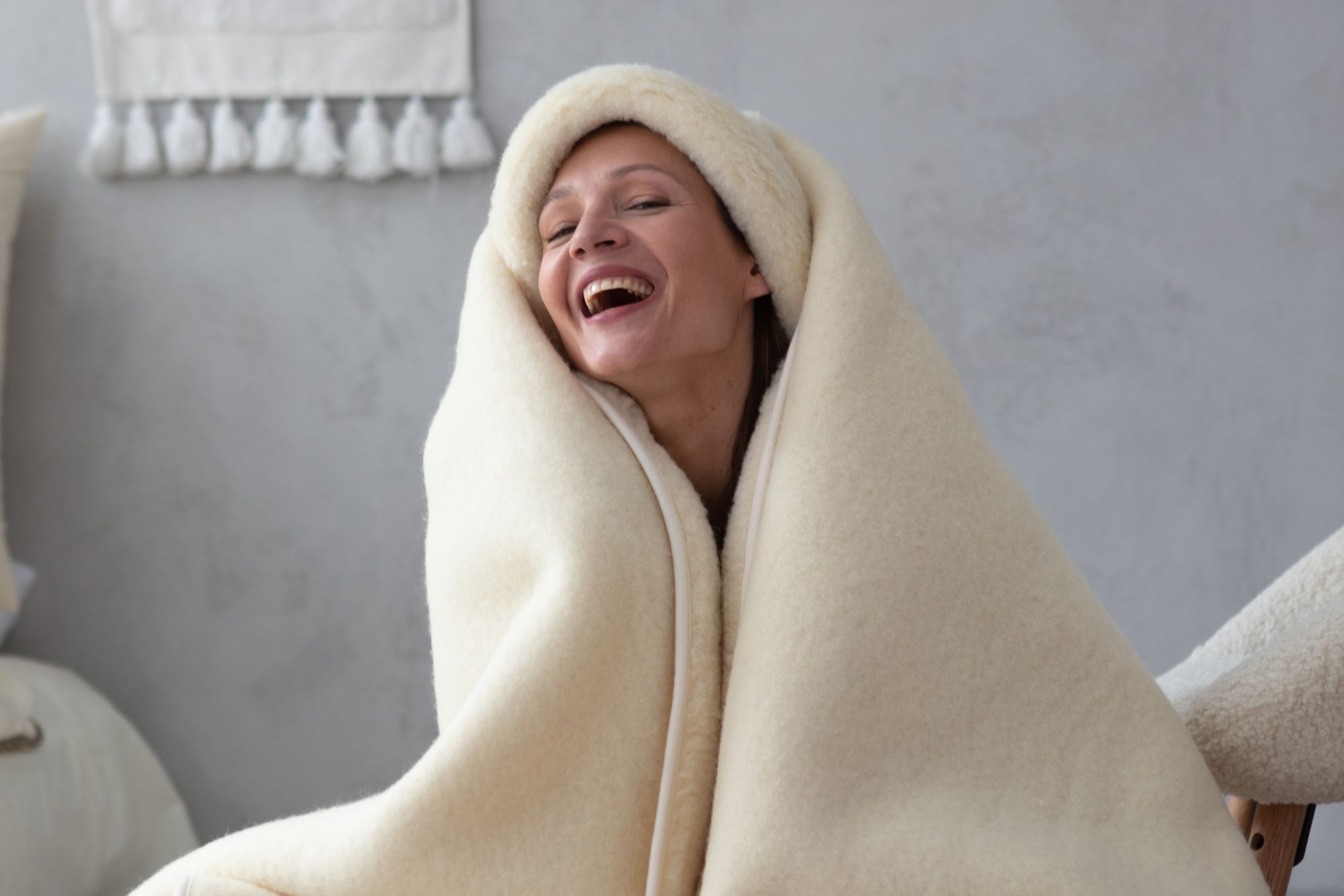 Kobieta owinięta w jasny, wełniany koc, uśmiecha się w przytulnym wnętrzu