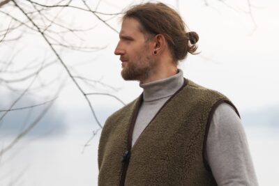 Mężczyzna w brązowej kamizelce wełnianej stoi na tle wody i drzew