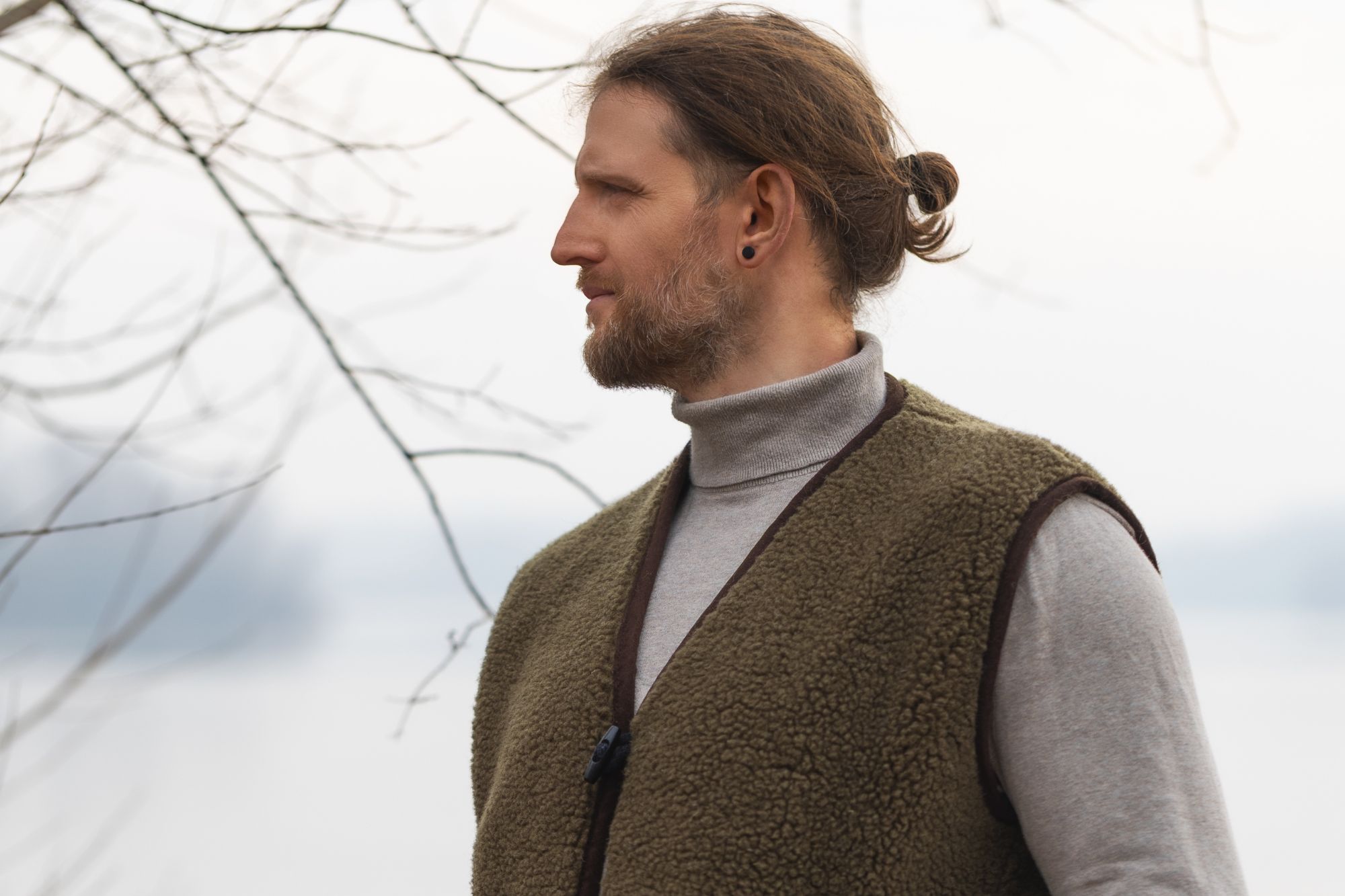 Mężczyzna w brązowej kamizelce wełnianej stoi na tle wody i drzew