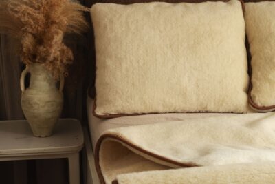 Poduszki w kolorze jasnego beżu i kołdra wełniana w tym samym odcieniu na łóżku obok wazonu z suszonymi kwiatami