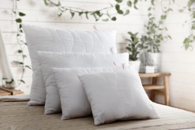 Cztery białe poduszki o różnej wielkości na łóżku, w tle rośliny i drewniany stół