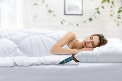 Kobieta śpiąca na białej poduszce, pod białą kołdrą, w jasnym wnętrzu z roślinami w tle