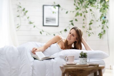 Kobieta leży na łóżku z białą kołdrą, czyta książkę w jasnym wnętrzu z roślinami